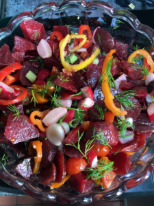 A vibrant beetroot salad in a glass serving bowl, featuring jewel-toned chunks of beetroot, sliced yellow and orange peppers, radishes, tomatoes, and sprigs of fresh dill. A riot of colour and care, prepared for a chavurah supper.