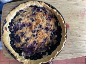 A rustic blueberry tart with a golden-brown crust and bubbling purple filling, just out of the oven. Slightly uneven, clearly homemade, and bursting with summer berries and soft cheese filling.