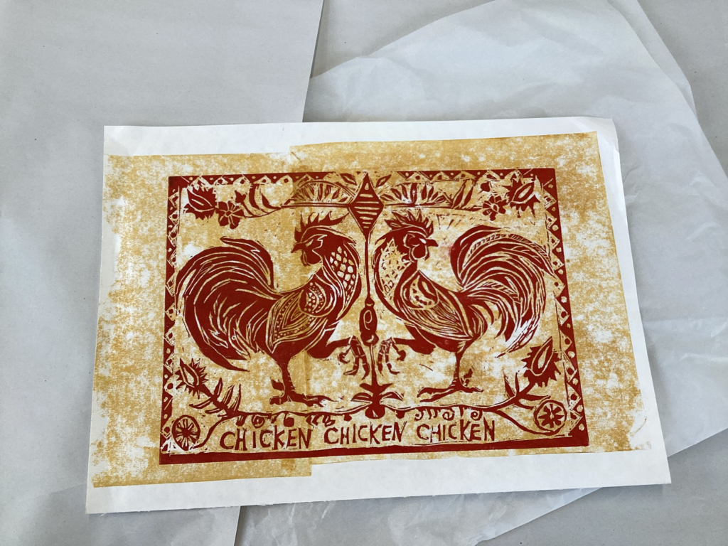A hand-carved lino print of two stylised chickens facing each other in mirror image, framed by folk-art inspired patterns and the repeated word “CHICKEN” at the bottom. Printed in red and ochre, bold and whimsical.
